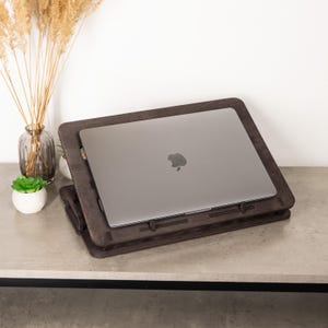 May include: A dark brown wooden laptop stand with a grey laptop on top. The stand is angled for ergonomic use and has a lower platform. Decorative items, including a vase with dried plants and a small succulent, are in the background.
