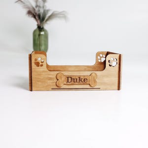 May include: A light brown wooden pet bed frame featuring a low-profile rectangular design with cut-out paw prints on the side panels and a central bone-shaped plaque engraved with the name Duke in a dark brown font on the front panel.