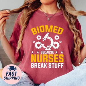 May include: Maroon t-shirt with the words "BIOMED BECAUSE NURSES BREAK STUFF" in yellow and white. The design includes a microscope and gears. A fast shipping logo is in the bottom left corner.