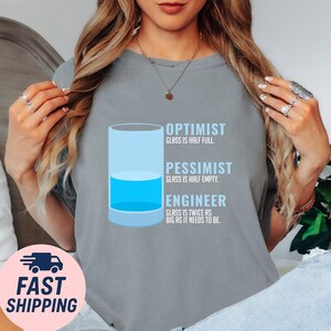 May include: Gray t-shirt with a graphic of a glass half full of blue liquid. Text reads: "Optimist: Glass is half full. Pessimist: Glass is half empty. Engineer: Glass is twice as big as it needs to be."