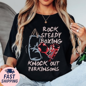 May include: Black t-shirt with the text "Rock Steady Boxing, Knock Out Parkinsons". The shirt features a graphic of a boxing bag and red boxing gloves. The shirt is a casual wear item.