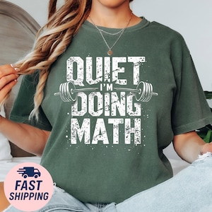 May include: A dark green t-shirt with the text "QUIET I'M DOING MATH" in a distressed white font. The design includes a barbell graphic. The shirt is a casual style, suitable for everyday wear.
