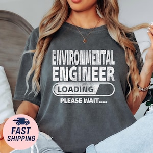 May include: Dark gray t-shirt with the text "ENVIRONMENTAL ENGINEER LOADING PLEASE WAIT....." in white. The shirt has a casual, slightly worn look.