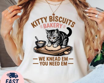 May include: Cream-colored t-shirt featuring a cartoon cat kneading dough on a wooden board. The text "KITTY BISCUITS BAKERY" arches above the cat, with "WE KNEAD EM - YOU NEED EM" below. A small bowl and spoon are also depicted.