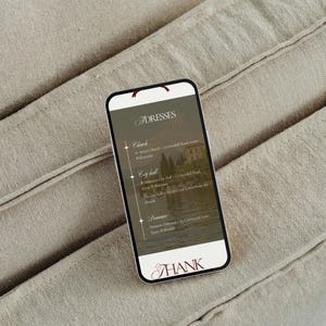 May include: A smartphone displaying a list of locations for dresses, including a church, city hall, and a domaine. The screen has a scenic background and the word "THANK" at the bottom. The phone rests on a beige fabric.
