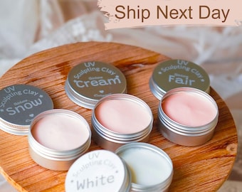 May include: A wooden tray holds several small, open metal tins of sculpting clay in various shades of pink and white. The lids are labeled with the words "Snow", "Cream", "Fair", and "White".