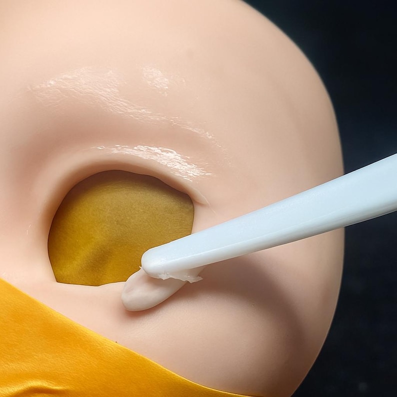 May include: Close-up of a light-skinned mannequin head with a large, open eye socket. A white tool applies a small amount of white substance to the edge of the eye socket. The eye socket is filled with a mustard-yellow material.