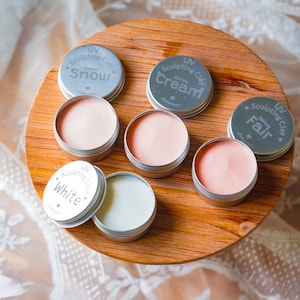 May include: A collection of small, round metal tins filled with sculpting clay in various shades of pink and white, arranged on a wooden surface. The lids are labeled with the names "Snow", "Cream", "Fair", and "White".