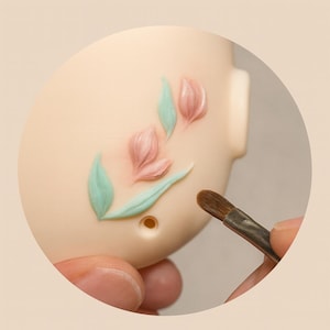 May include: Close-up of a light beige figurine being decorated with pink and green floral accents. A small paintbrush is held near the figurine, suggesting the process of adding details to the design. The figurine has a smooth surface.