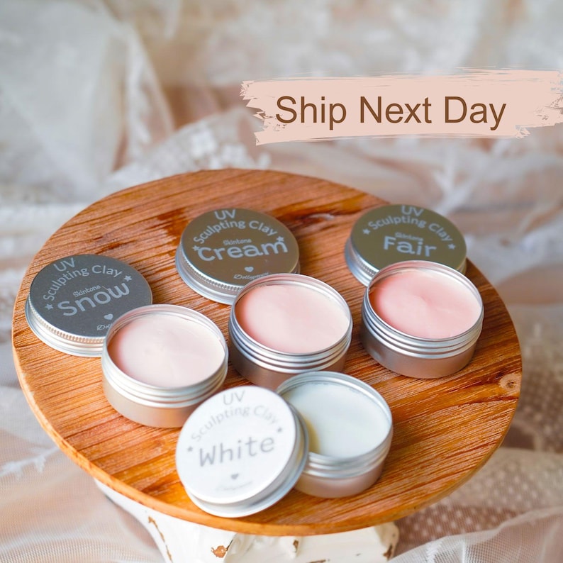 May include: A wooden tray holds several small, open metal tins of sculpting clay in various shades of pink and white. The lids are labeled with the words "Snow", "Cream", "Fair", and "White".