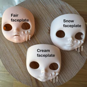 May include: Three doll faceplates in varying shades of skin tone: Fair, Snow, and Cream. Each faceplate has large eye sockets, a small nose, and a defined mouth. The text "Fair faceplate", "Snow faceplate", and "Cream faceplate" are printed on each faceplate.