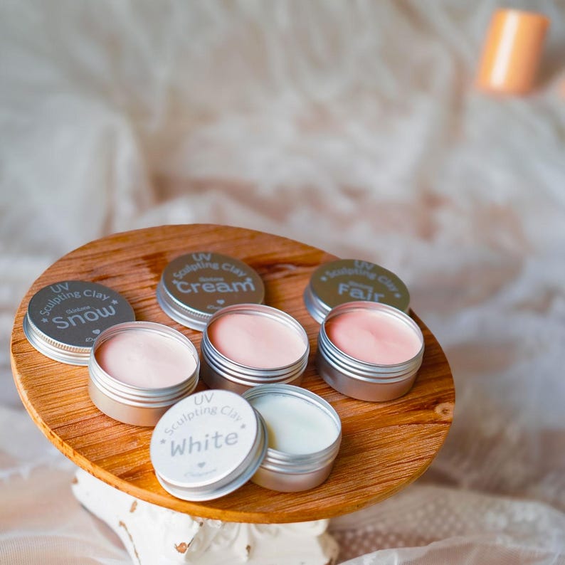 May include: A wooden serving tray holds several small, round, silver tins of UV Sculpting Clay. The tins contain various shades of cream and white clay. The lids are labeled with the names "Snow", "Cream", "Fair", and "White".