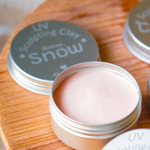 May include: A close-up of a small, open silver tin filled with a light pink substance, likely a cosmetic product. The lid, with the text "UV Sculpting Clay Snow", is next to the open tin. The background is a wooden surface.