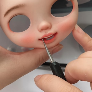 May include: A close-up of a doll's face with painted features, including rosy cheeks, freckles, and open eye sockets. A tool is used to paint the doll's lips, which are a coral color. The doll's face is a light peach color.