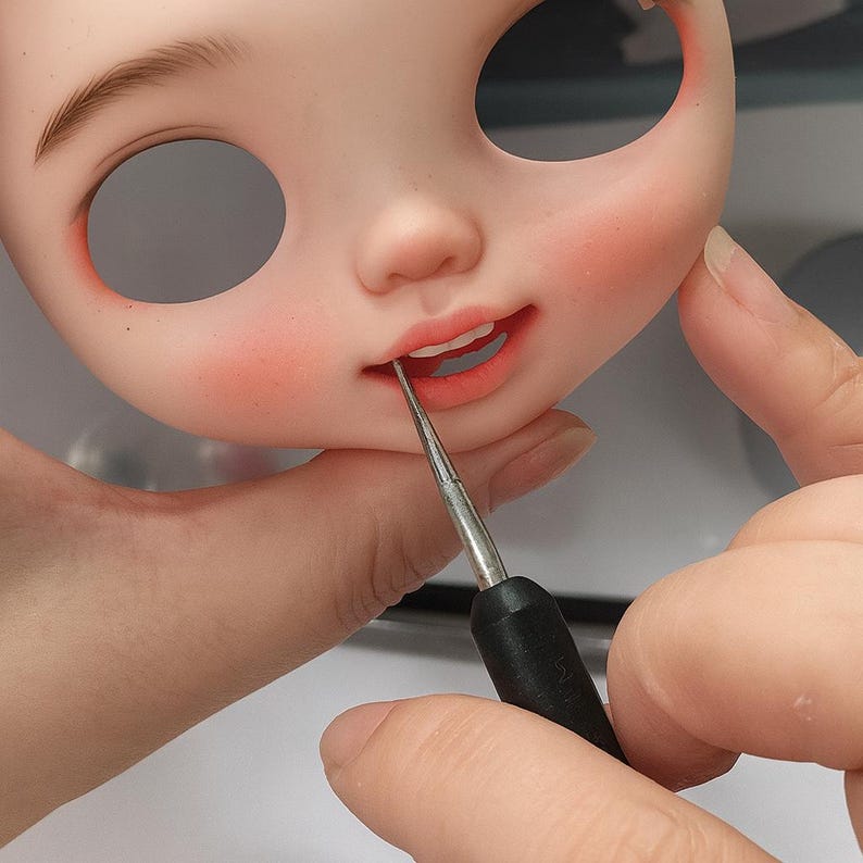 May include: A close-up of a doll's face with painted features, including rosy cheeks, freckles, and open eye sockets. A tool is used to paint the doll's lips, which are a coral color. The doll's face is a light peach color.