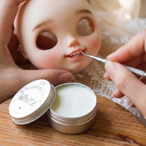 May include: A doll's head with painted features, including rosy cheeks and detailed lips. A small metal container of white sculpting clay is open next to the doll's face. The lid of the container reads "UV Sculpting Clay White."