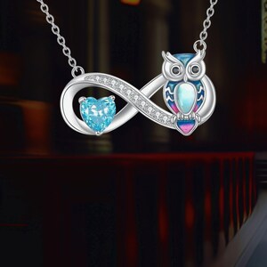 Könnte beinhalten: Silberkette mit Eulen- und Unendlichkeitszeichen-Design. Die Eule hat blaue, rosa und weiße Emaille-Details. Ein herzförmiger, hellblauer Edelstein ist in das Unendlichkeitszeichen eingesetzt. Die Halskette hängt an einer filigranen Kette.