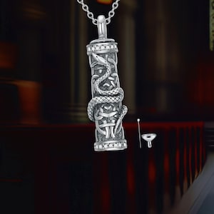May include: Silver pendant necklace with a cylindrical design, featuring a snake wrapped around it. The pendant has Celtic knotwork and is decorated with small, clear stones. A small funnel and tool are included.