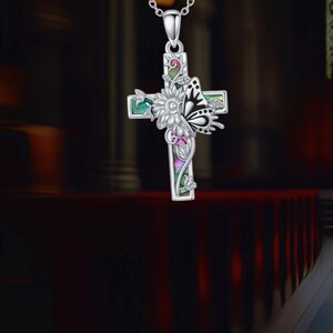 Könnte beinhalten: Eine silberfarbene Anhängerkette mit einem Kreuzdesign. Das Kreuz ist mit einem Schmetterling, einer Blume und farbenfrohen, irisierenden Akzenten verziert. Der Schmetterling hat schwarz-weiße Details. Die Halskette hängt an einer filigranen Silberkette.