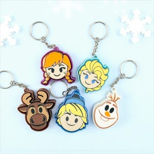May include: A collection of keychains featuring characters from a popular animated film. The keychains include Anna, Elsa, Kristoff, Olaf, and Sven. Each keychain has a metal ring and chain, with the character's face as the main design element. The keychains are colorful and made of a rubbery material.