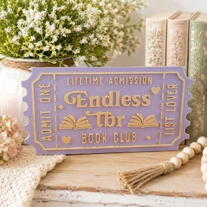 Könnte beinhalten: Ein lilafarbenes Schild in Form eines Tickets mit den Worten "Endless TBR Book Club" in goldfarbenen Buchstaben. Das Schild enthält den Text "Lifetime Admission" und "List Lover". Dekorative Elemente sind offene Bücher und Herzen.