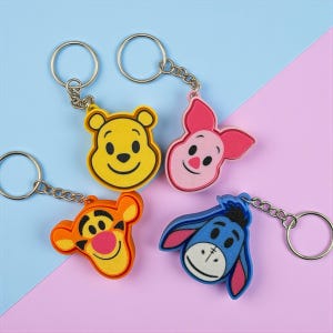 Op de afbeelding: Vier sleutelhangers met de personages Winnie de Poeh, Knorretje, Tijgertje en Iejoor. Elke sleutelhanger heeft een zilverkleurige ketting en ring. De karakters zijn geel, roze, oranje en blauw, met zwarte en roze accenten. De achtergrond is diagonaal verdeeld, met blauwe en roze secties.