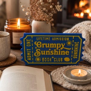 May include: A blue and yellow sign that reads "Lifetime Admission Grumpy Sunshine Book Club" sits on a wooden surface. A lit candle, a cup of tea, and an open book are also on the table, creating a cozy reading atmosphere.
