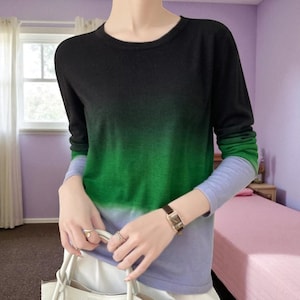 Puede incluir: Una camisa de manga larga con degradado de color. La parte superior pasa de negro en los hombros a verde en el medio, y luego a azul en las muñecas. La camisa tiene un cuello redondo.