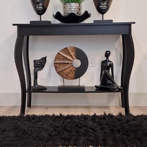 Puede incluir: Una consola negra con objetos decorativos. Dos máscaras de madera tallada flanquean una planta en maceta. La balda inferior contiene una escultura de rostro, una obra de arte circular y una figura sentada. Una alfombra negra está en primer plano.