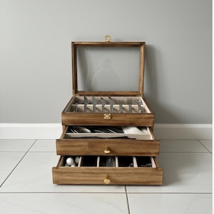 Puede incluir: Un cofre de cubiertos de madera con tres cajones y una tapa de cristal. El cofre está abierto, revelando cubiertos cuidadosamente organizados. Los cajones tienen pomos de latón. El cofre es de madera clara y está sobre un suelo de baldosas blancas.