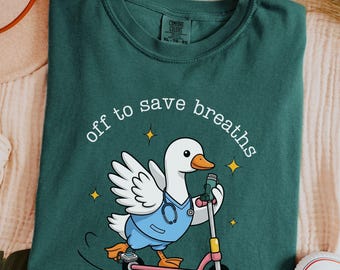 May include: Blue spruce t-shirt with a cartoon duck riding a pink scooter. The duck is wearing a blue uniform and stethoscope, with the text "off to save breaths". The shirt is next to a hat and white sneakers.
