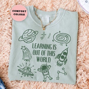 May include: Sage green t-shirt with a space-themed graphic. The design includes a planet, shooting star, rocket, UFO, astronaut, and the text "LEARNING IS OUT OF THIS WORLD". A gold necklace and white sneakers are also visible.