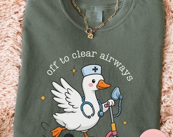May include: Moss green t-shirt with a cartoon goose wearing a nurse's hat and stethoscope, riding a pink scooter. The text "off to clear airways" is arched above the goose. A gold chain necklace is visible. The shirt is labeled "Comfort Colors".