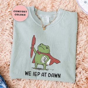 May include: A light green t-shirt featuring a cartoon frog superhero holding a red pen and a notepad labeled "IEP". The text "WE IEP AT DAWN" is printed below the frog. A gold necklace is visible.