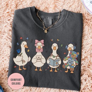 May include: Dark gray t-shirt featuring a whimsical illustration of four cartoon geese, each with unique accessories. One goose holds a rose, another wears a pink bow, a third reads a book, and the last one holds a teddy bear. The shirt has the words "Comfort Colors" and "Pepper".