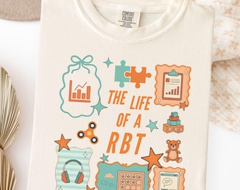 May include: Ivory-colored t-shirt with the text "THE LIFE OF A RBT" and various graphic elements, including puzzle pieces, a graph, a teddy bear, and headphones. The shirt is made of a soft material.