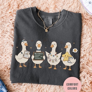 May include: Dark gray t-shirt featuring four cartoon ducks, each holding items like a chart, a sign with text, and a star. The shirt has a crew neck and a gold necklace. The text on the sign reads "ABCs of Behavior".