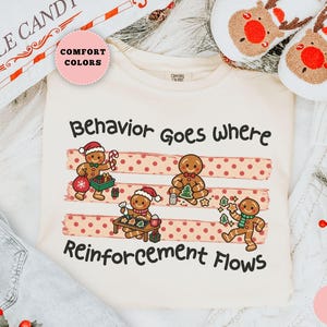 May include: An ivory t-shirt featuring a graphic of four gingerbread people in various holiday activities. The text reads Behavior Goes Where Reinforcement Flows. The design includes three horizontal red polka-dot brushstroke stripes.