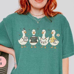 May include: Light green t-shirt featuring a whimsical design of four cartoon ducks. Each duck holds an item, including a star, a document, and a sign that reads "ABCs of Behavior". The shirt's color is light green.