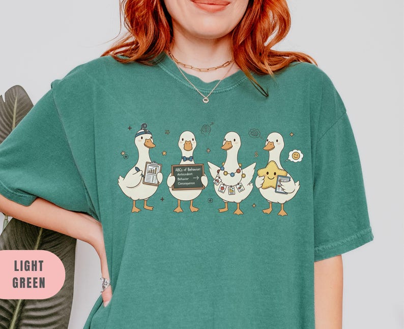May include: Light green t-shirt featuring a whimsical design of four cartoon ducks. Each duck holds an item, including a star, a document, and a sign that reads "ABCs of Behavior". The shirt's color is light green.