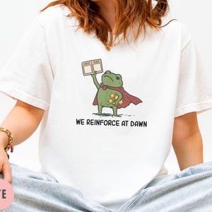 May include: White t-shirt featuring a cartoon frog superhero with a red cape and stars. The frog holds a sign that says "FIRST THEN". The text "WE REINFORCE AT DAWN" is printed below the frog.