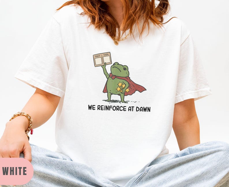 May include: White t-shirt featuring a cartoon frog superhero with a red cape and stars. The frog holds a sign that says "FIRST THEN". The text "WE REINFORCE AT DAWN" is printed below the frog.