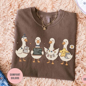 May include: Espresso-colored t-shirt featuring four cartoon ducks, each with unique accessories and holding items like a chart and a star. The shirt has the text "Comfort Colors" and "Espresso" on it. A gold necklace is visible.