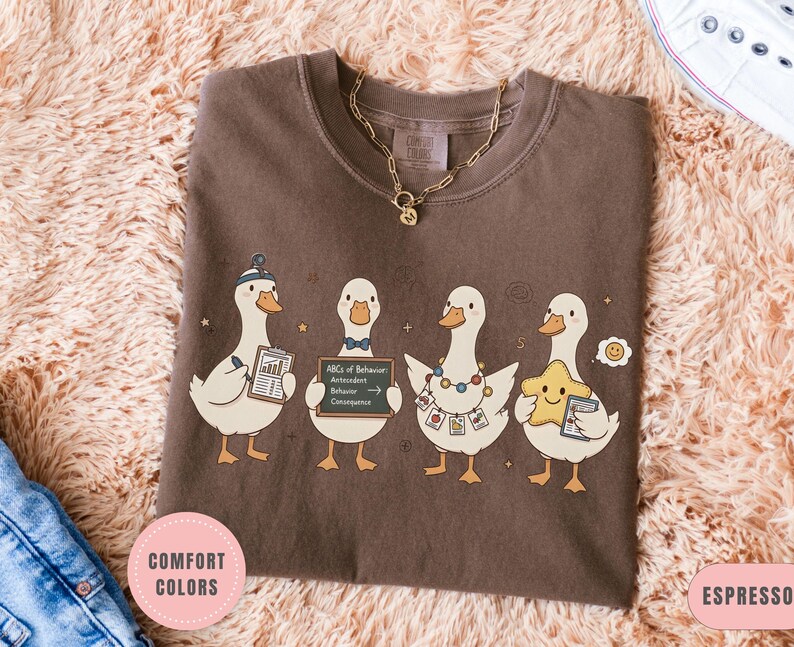 May include: Espresso-colored t-shirt featuring four cartoon ducks, each with unique accessories and holding items like a chart and a star. The shirt has the text "Comfort Colors" and "Espresso" on it. A gold necklace is visible.