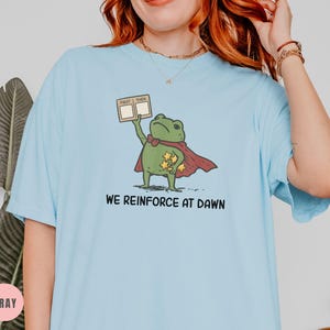May include: Light blue t-shirt featuring a cartoon frog superhero with a red cape and gold stars. The frog holds a sign that says "First Then". The text "We Reinforce at Dawn" is printed below the frog.