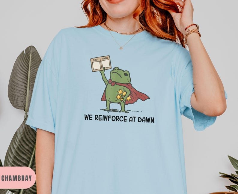 May include: Light blue t-shirt featuring a cartoon frog superhero with a red cape and gold stars. The frog holds a sign that says "First Then". The text "We Reinforce at Dawn" is printed below the frog.