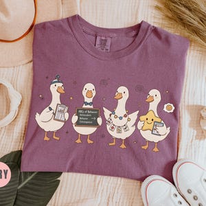 May include: Berry-colored t-shirt featuring four cartoon ducks, each holding items like a clipboard and a sign that reads "ABCs of Behavior." The ducks are adorned with accessories, including a hat and a star.