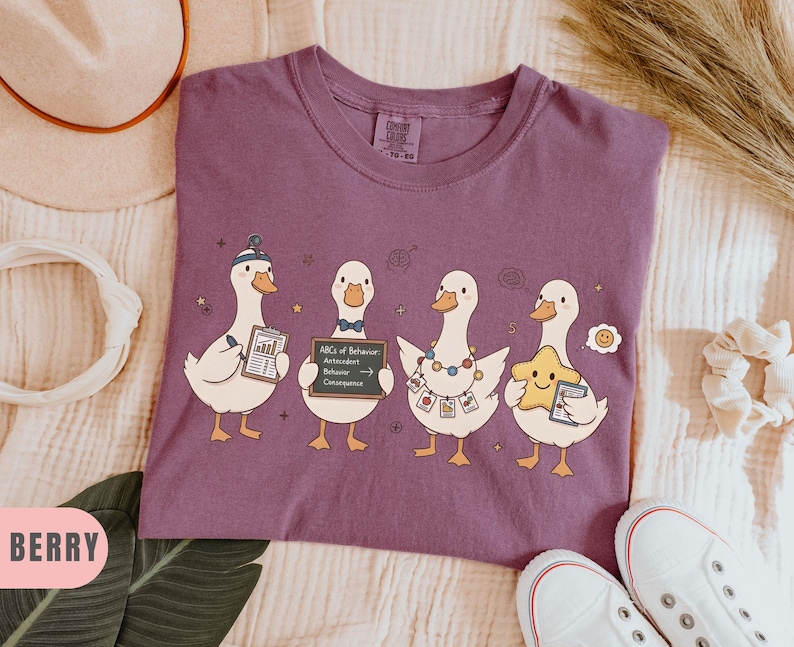 May include: Berry-colored t-shirt featuring four cartoon ducks, each holding items like a clipboard and a sign that reads "ABCs of Behavior." The ducks are adorned with accessories, including a hat and a star.
