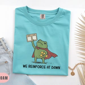 May include: Sea foam green t-shirt featuring a cartoon frog superhero with a red cape and gold stars. The frog holds a sign that says "FIRST THEN". The text "WE REINFORCE AT DAWN" is printed below the frog. The shirt is folded.
