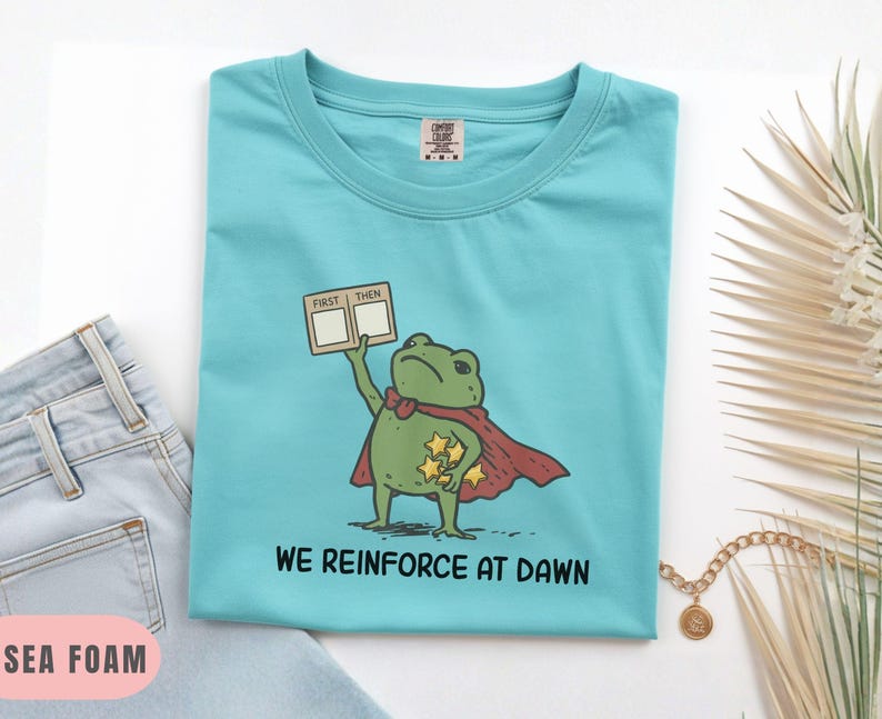May include: Sea foam green t-shirt featuring a cartoon frog superhero with a red cape and gold stars. The frog holds a sign that says "FIRST THEN". The text "WE REINFORCE AT DAWN" is printed below the frog. The shirt is folded.